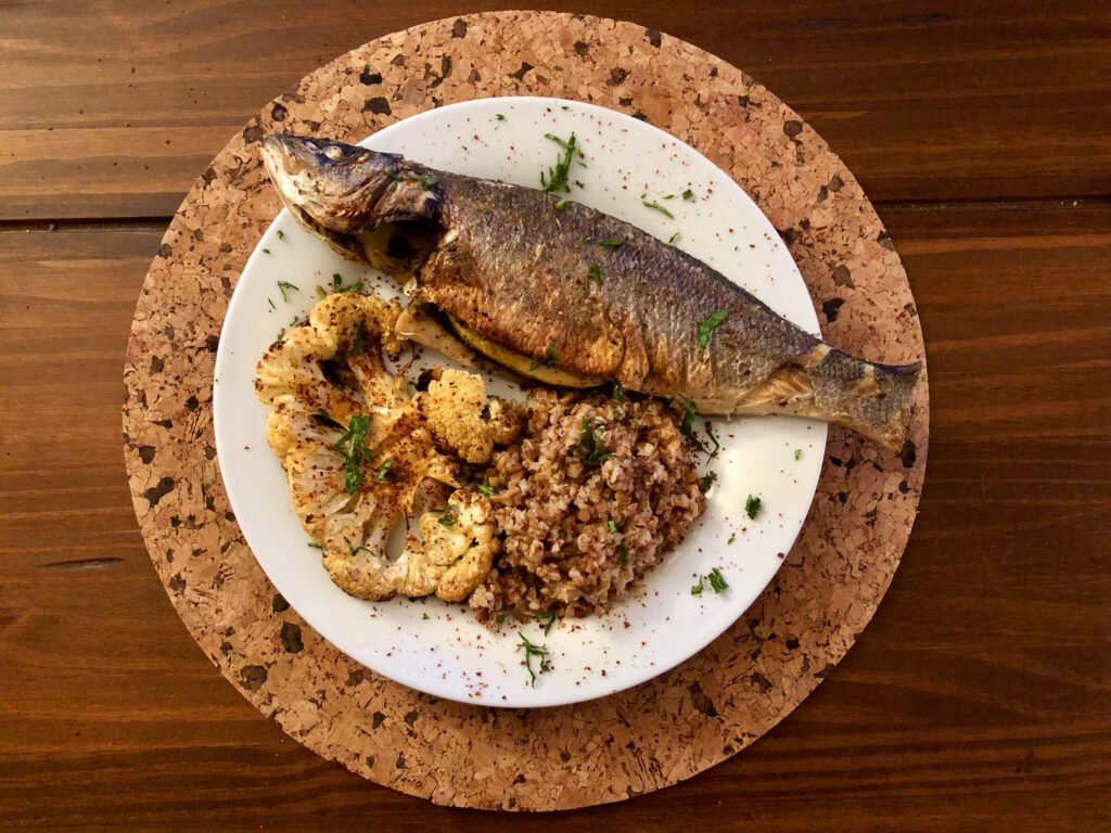Grilled whole fish with roasted cauliflower and seasoned grains, garnished with herbs on a white plate over a cork trivet and wooden table.
