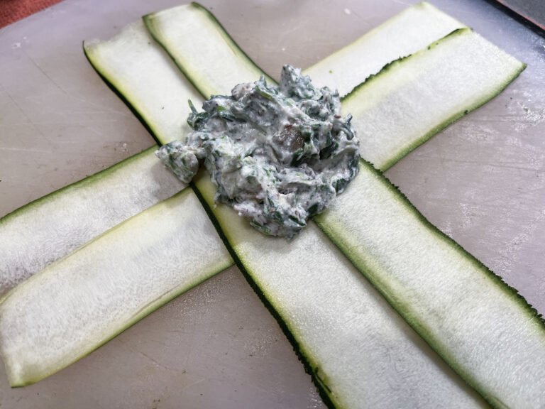 Thin zucchini ribbons cross on a cutting board beneath a mound of creamy herb filling, arranged like a rustic vegetable parcel in a calm kitchen setting.