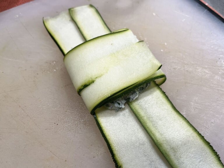 Pale green zucchini ribbons are folded around a small dark filling on a cutting board, forming a neat, fresh-looking vegetable parcel.