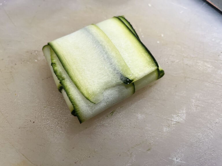 A neat square parcel of overlapping pale-green zucchini ribbons, edged with dark green peel, rests on a cutting board, suggesting fresh, careful preparation.