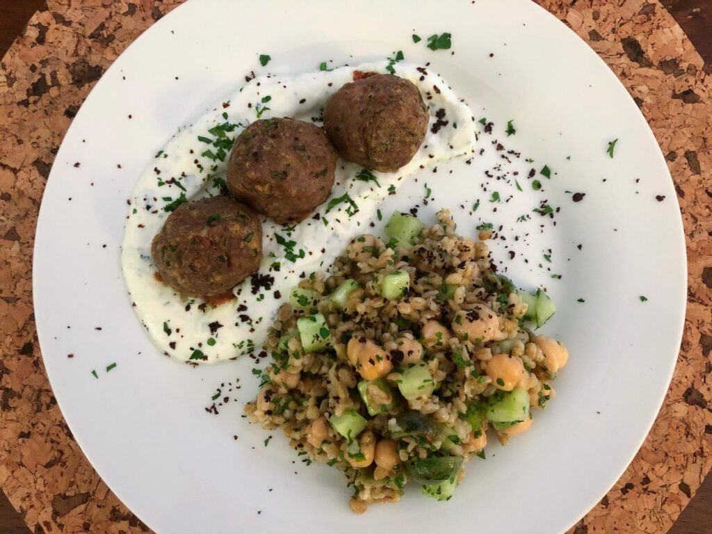 A white plate holds three browned falafel balls on herb-flecked yogurt beside a hearty grain salad with chickpeas and cucumber, presented on a cork table.