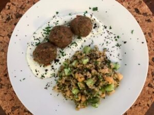 A white plate holds three browned falafel balls on herb-flecked yogurt beside a hearty grain salad with chickpeas and cucumber, presented on a cork table.