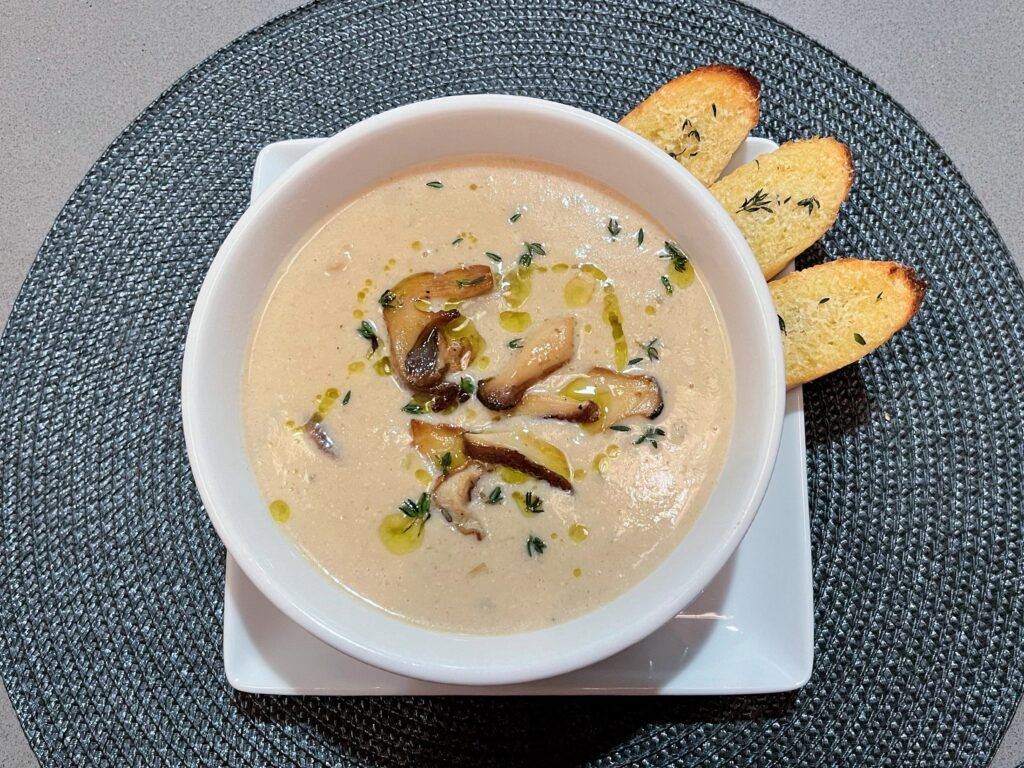 A white bowl of creamy mushroom soup, drizzled with olive oil and herbs, sits on a square plate with three toasted bread slices; warm, inviting presentation.