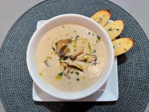 A white bowl of creamy mushroom soup, drizzled with olive oil and herbs, sits on a square plate with three toasted bread slices; warm, inviting presentation.