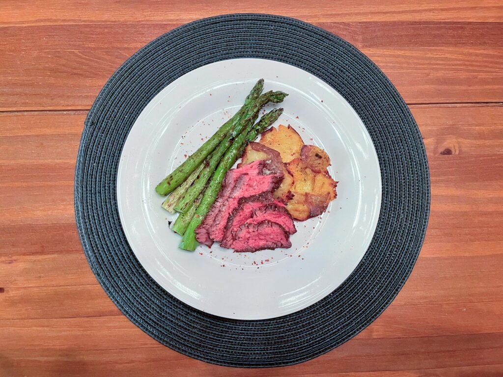 A white plate on a dark woven placemat holds sliced pink-centered roast beef, grilled asparagus, and golden roasted potatoes on a wooden table, presenting a warm meal.