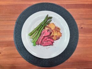 A white plate on a dark woven placemat holds sliced pink-centered roast beef, grilled asparagus, and golden roasted potatoes on a wooden table, presenting a warm meal.
