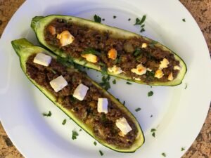 Two baked zucchini halves on a white plate, filled with browned minced meat and topped with melted cheese and chopped herbs; the dish looks hearty and appetizing.