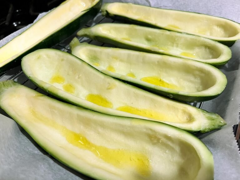 Halved zucchini boats with their centers scooped out and drizzled with oil rest on a baking tray, ready to be stuffed or roasted; fresh and inviting.