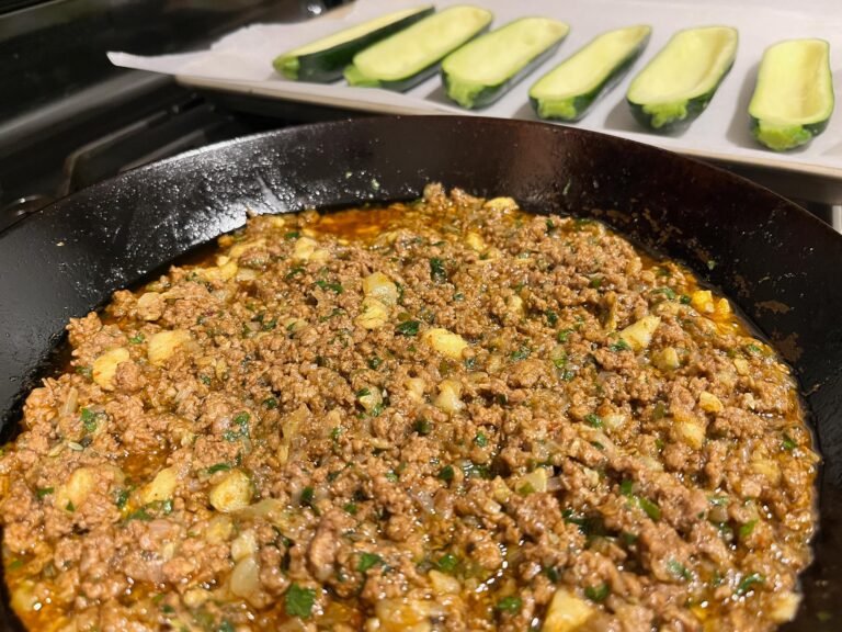 A black skillet holds a browned ground-meat and potato mixture flecked with herbs, while sliced zucchini rests on a tray behind it, ready to cook.
