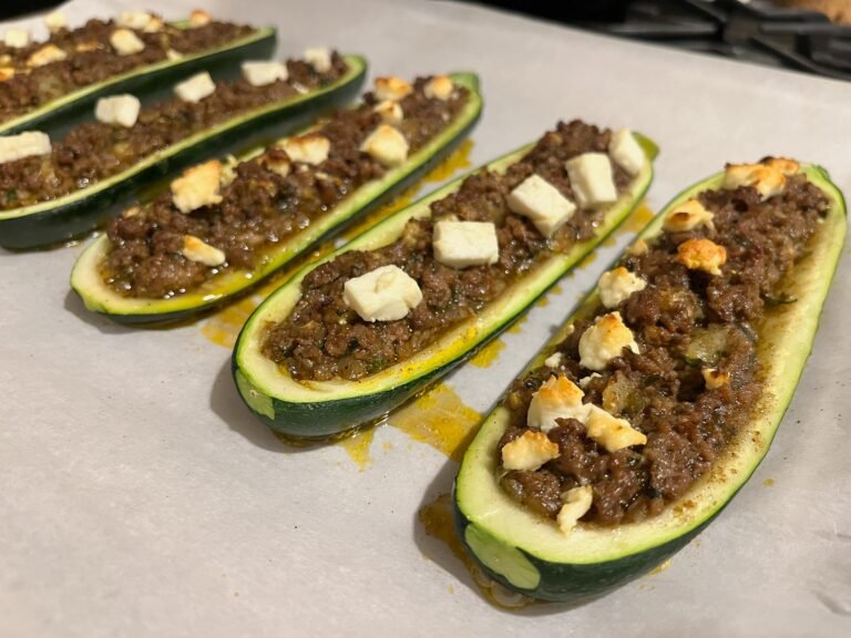 Several zucchini boats are filled with browned minced meat and cheese, arranged on a baking tray; the golden, hearty dish looks warm and appetizing.